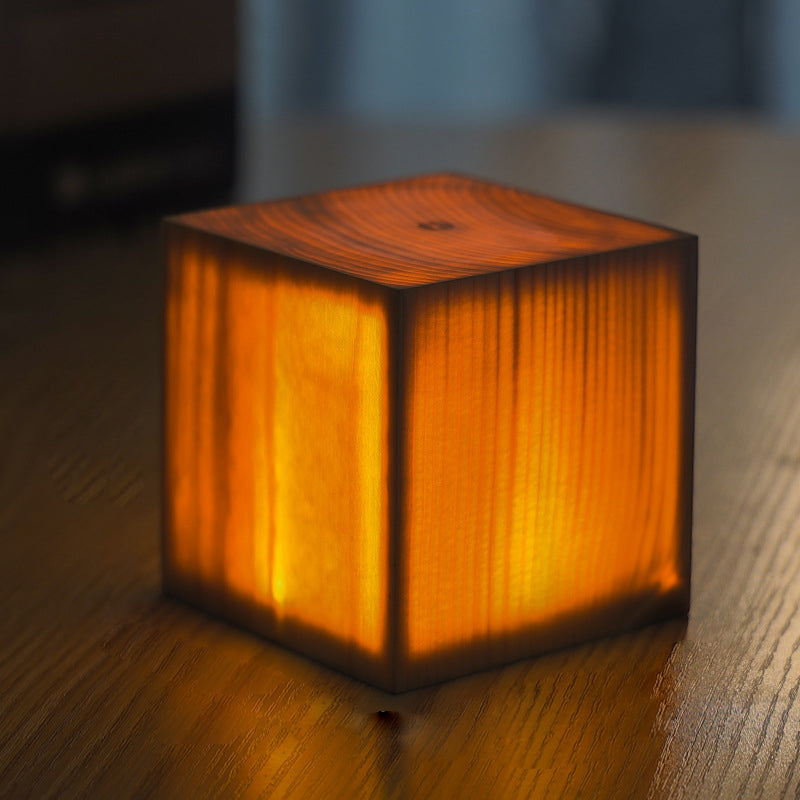 Square wood lamp in cylindrical shape, glowing warmly on a wooden surface.